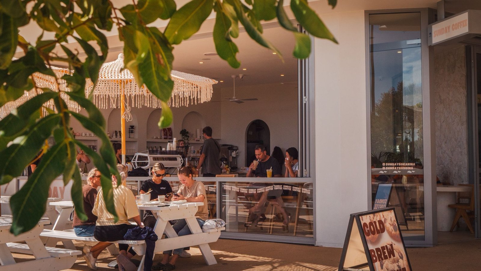 Outdoor café scene at Sunday Coffee in Bokarina, featuring patrons enjoying drinks at white picnic tables under a decorative umbrella, with a focus on the café's inviting atmosphere and coffee offerings.
