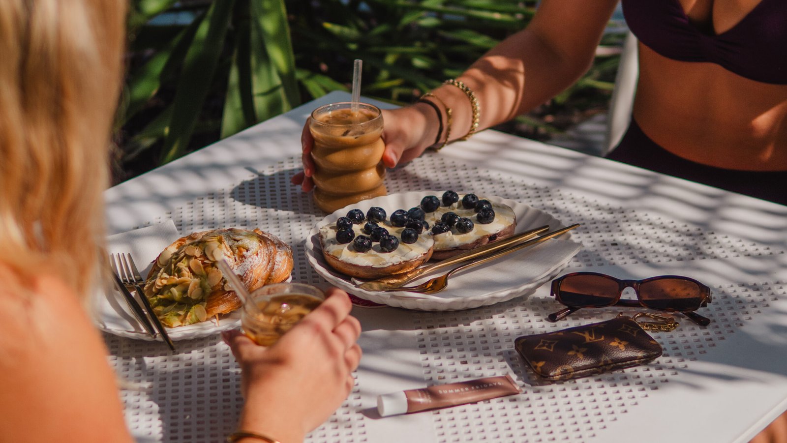 Two women enjoying made-to-order bagels topped with cream cheese and blueberries, accompanied by iced coffee and pastries, on a sunlit table with greenery in the background.